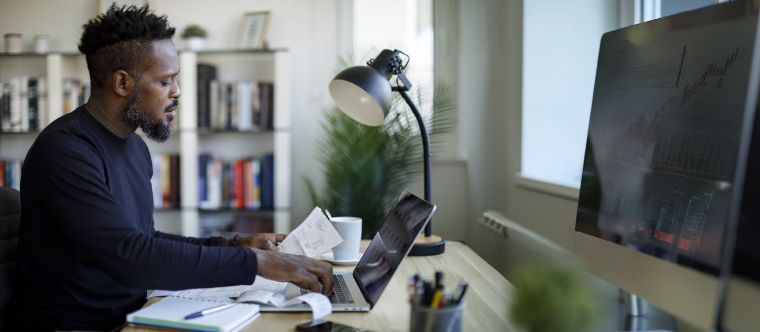 Image of a man sitting in a home office type setting, with a laptop and various papers.