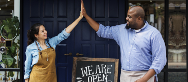 Image of two people giving each other a high-five outside of a business with a "We are open" sign.