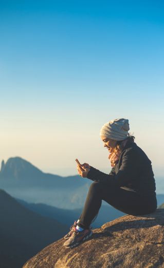 Image of woman on a large rock in front of mountains, holding a cell phone.