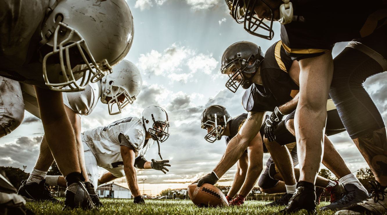 Image of football players head-to-head preparing for a play on a green football field with a lightly clouded sky in the background. One team has white uniforms and helmets, the other team has black uniforms and helmets.