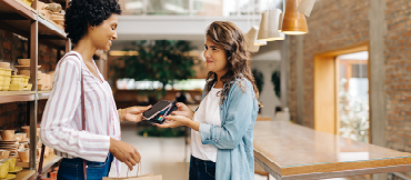 Image of two women inside a building with one using a phone to pay on a merchant services terminal.