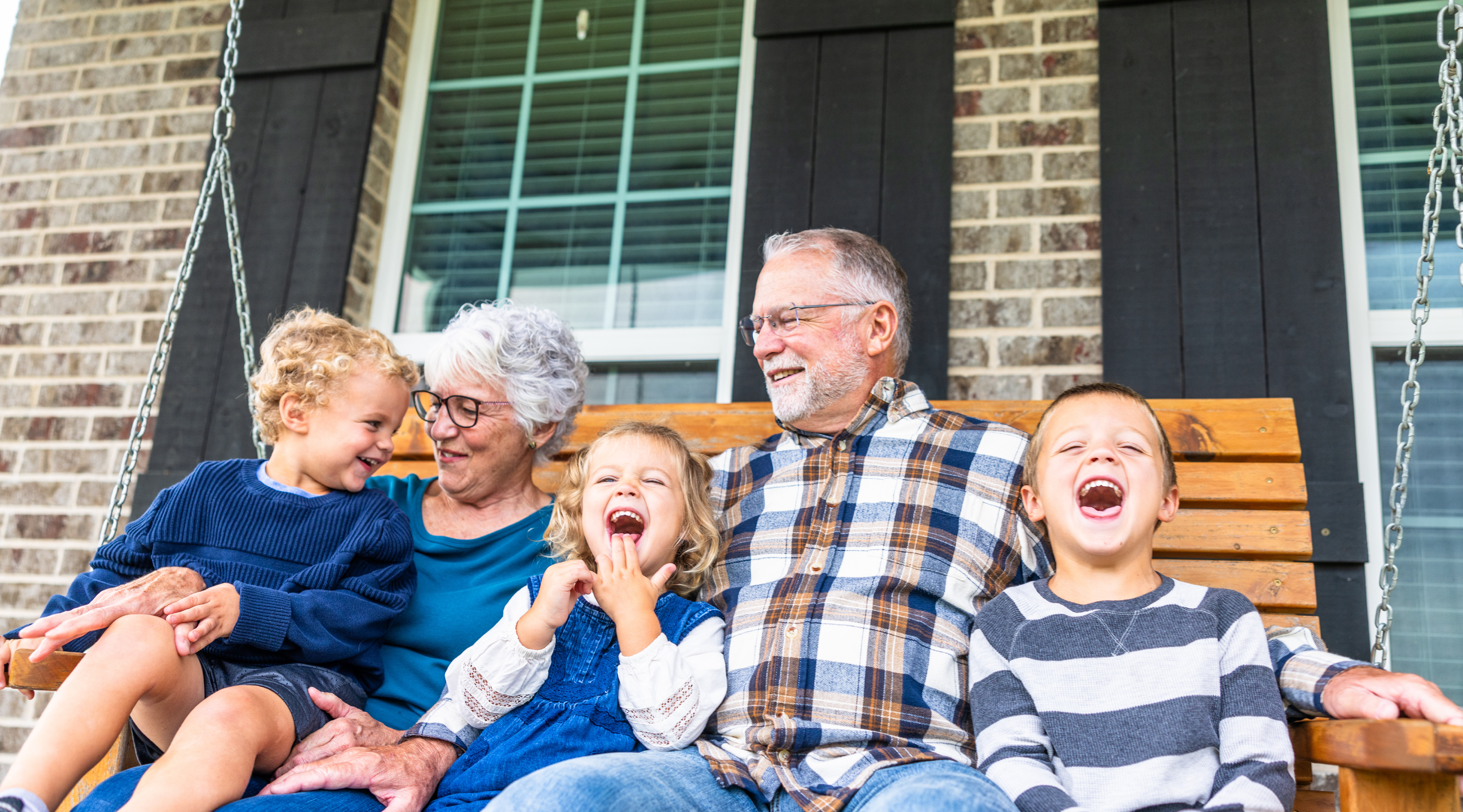 Image of 3 young children and two adults sitting on a wooden porch swing in front of a brick house with windows and black shutters. All individuals appear to be smiling and happy.